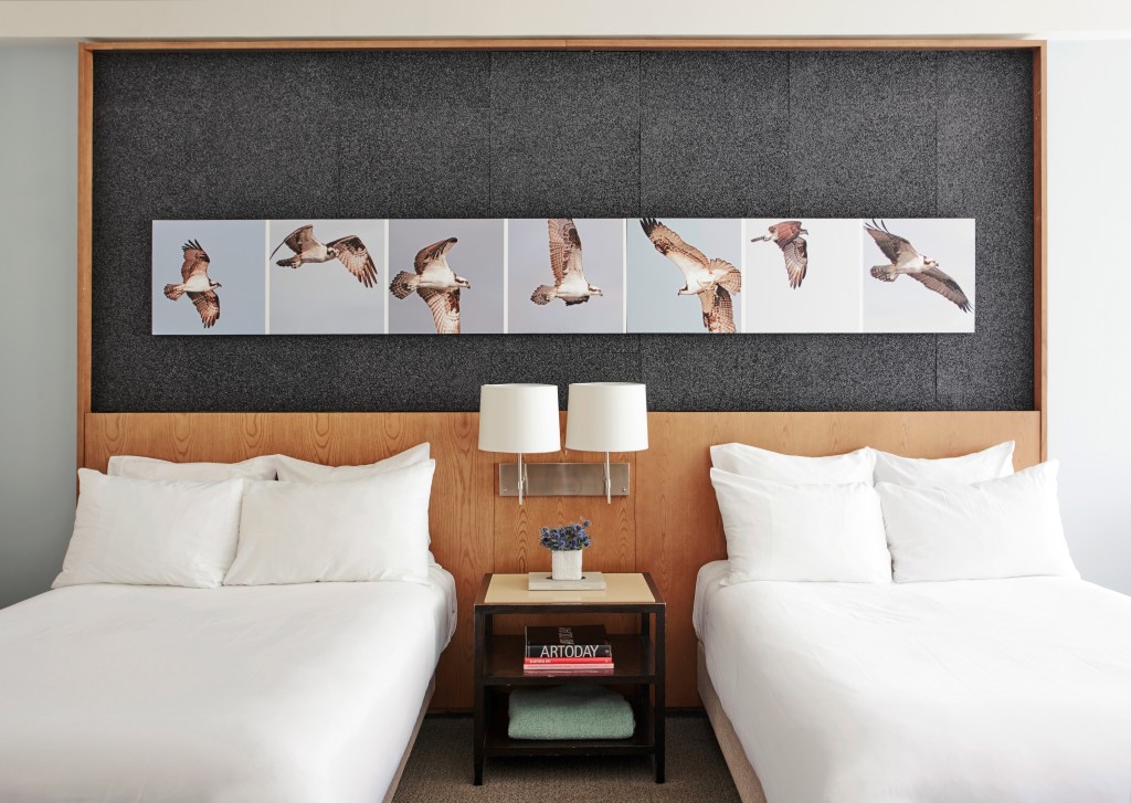 A hotel room with two queen beds and a nightstand in between with lamps and artwork above the bed
