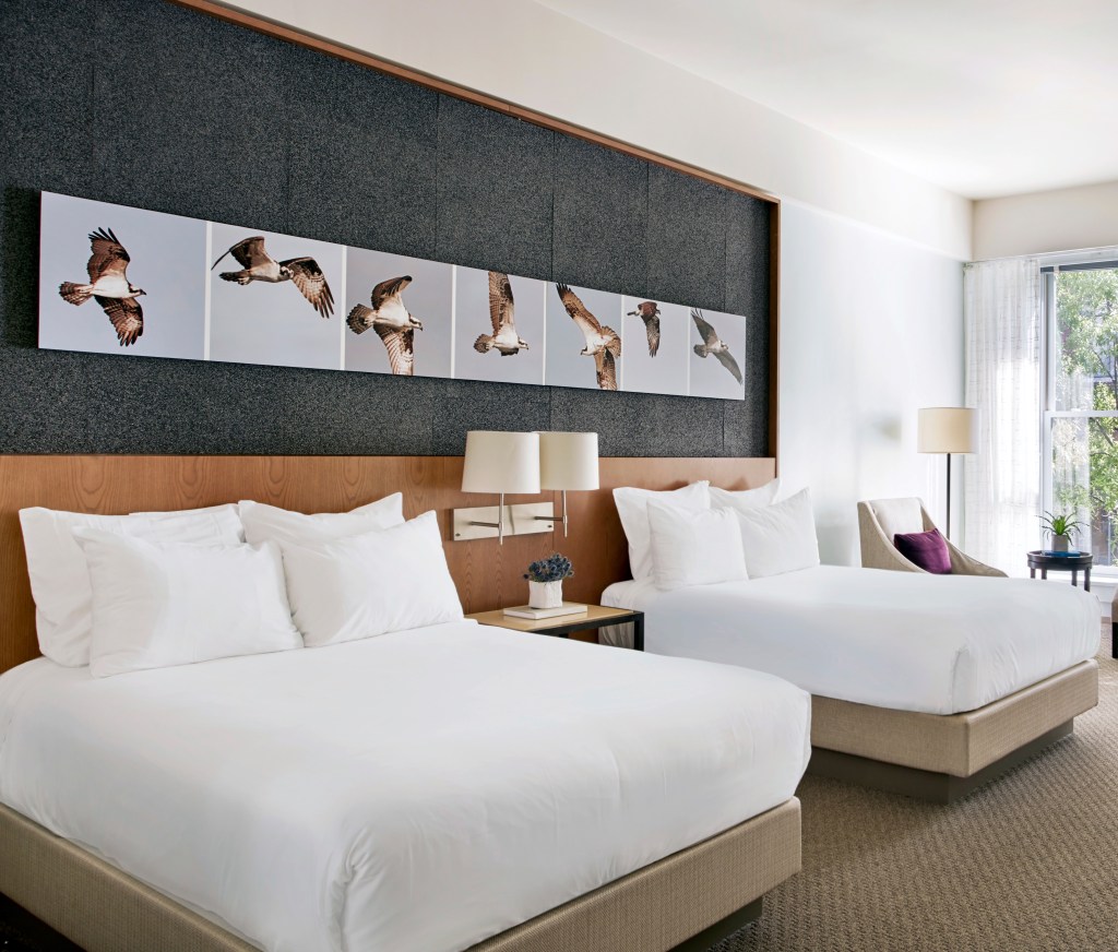 A hotel room with two queen beds, nightstand, and side chair in a spacious room with large windows