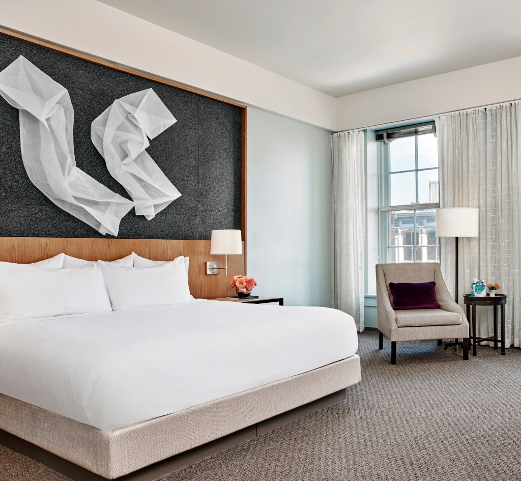 A hotel room with one king bed, chair with lamp and side table in a spacious room with large windows