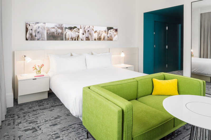 Large white bed in hotel room with green sofa at foot of bed and small white bistro table, two nightstands are on either side of the bed